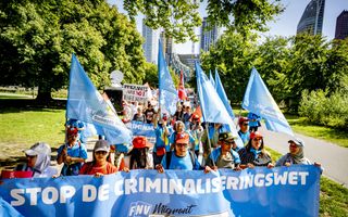 Een stoet mensen loopt op een pad in een park met lichtblauwe vlaggen en allerlei protestborden en vooraan een rij die een groot lichtblauw spandoek vasthoudt.