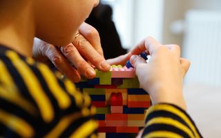 Een jongetje draagt een trui met gele en zwarte strepen. Hij bouwt een toren van Lego. Een volwassene helpt mee.