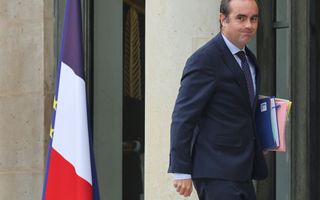 Een man in pak poseert bij de ingang van een paleis. Onder zijn armen draagt hij stapels papieren. Naast hem staan vlaggen. 