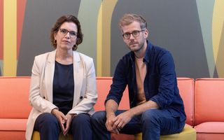 Een vrouw en een man zitten, ieder op een poef, naast elkaar in een studio en kijken, met serieuze blik, in de lens. 