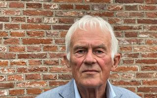 Portretfoto van oudere man met grijs haar. Hij draagt een blauwgrijs colbert op een lichtblauw overhemd. Hij staat tegen de achtergrond van een bakstenen muur.