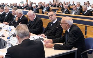 Mannen in zwarte pakken zitten aan vergadertafels; een van hen is aan het woord. Achter hen staan kerkbanken vol zittende mannen.
