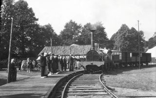 Zwart-witfoto van een oude stoomlocomotief met enkele rijtuigen die bij een houten perron stilstaat. Op het perron staan mensen in historische kledij en op klompen. Op de achtergrond zijn bomen te zien. Links staat een paard-en-wagen.