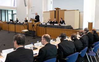 Predikanten in zwarte pakken zitten aan vergadertafels in een kerkgebouw.