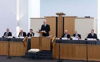 Zes mannen in zwarte pakken, vijf zittend en één staand, achter vergadertafels in een modern kerkgebouw.