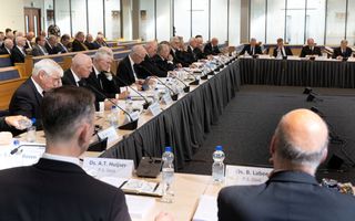 Zo’n twintig mannen in zwart pak zitten achter tafels in een modern kerkgebouw.