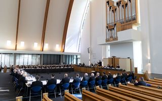 Veertig mannen in zwarte pakken zitten in een modern kerkgebouw op blauwe stoelen rond vergadertafels die in een vierkant zijn opgesteld.