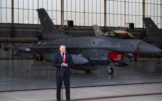 Man in formele kleding achter microfoon. Een gevechtsvliegtuig staat achter hem.