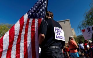 Een jongeman met op zijn rug een wit papier met de woorden Pray for America. Hij draagt een Amerikaanse vlag.