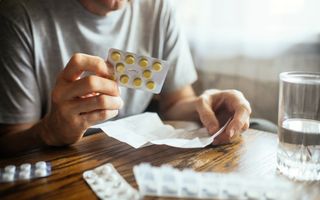 Een man in grijs T-shirt leest de bijsluiter van een medicijn. Rechts een glas water.