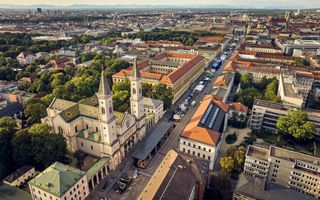 Luchtfoto van München waarop op een doorgaande weg allerlei stands te zien zijn van automerken. Links op de voorgrond een mooie kerk met twee torens, daarachter de skyline van München. 