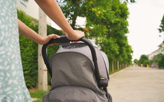 Deels zichtbare vrouw achter kinderwagen op voetpad met bomen erlangs.