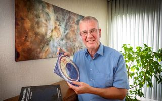 Man in woonkamer, leunend tegen een tafel met een boek.