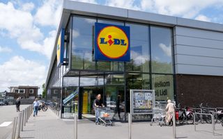 Glazen gevel van een vestiging van Lidl, met daarop groot het logo. Voor de winkel mensen, winkelwagens en fietsen. 