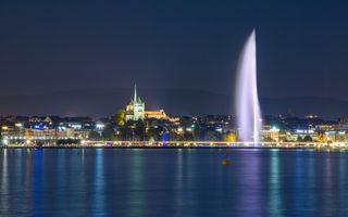 De Zwitserse stad Genève bij nacht, gezien vanaf het meer. Op de voorgrond een fontein. In het midden een verlichte kathedraal.