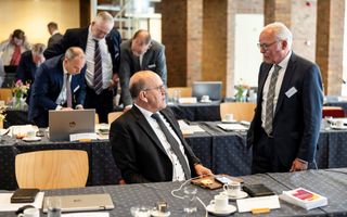 Lange vergadertafels, met daarachter mannen in donkere pakken, staand of zittend, met elkaar in gesprek. 