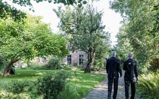 Twee mannen in zwarte pakken lopen naar een door groen omzoomde woning toe.