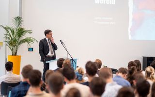 Man in donker pak staat achter een katheder en spreekt; er zitten tientallen jongeren in de zaal.