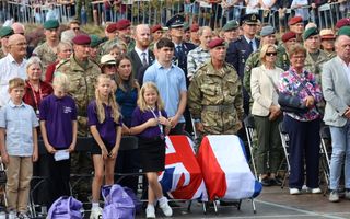 Publiek en soldaten staan op hun plaatsen. Over de twee lege stoelen zijn de vlaggen van het Verenigd Koninkrijk en Nederland gedrapeerd.