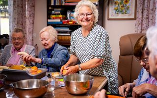 Een aantal mensen zitten rondom een tafel en schillen een banaan en een sinaasappel. Een van de vrouwen staat en kijkt lachend in de camera. 