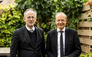 Dominee De Vries en dominee Brons, twee mannen met bril en zwart pak, in een tuin.