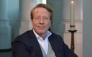 Predikant met bruin haar en bril zit voor kaars in kerk.