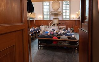 Kerkzaal met mensen in banken, gezien door twee openstaande houten deuren.