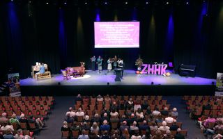 Een donkere zaal met tientallen bezoekers die een psalm zingen, begeleid door instrumentalisten op het podium.