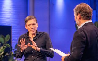 Man in zwart overhemd spreekt op een podium.