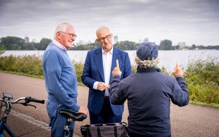 Man in gesprek met twee voorbijgangers. Hij luistert naar een man met een petje die met beide vingers in de lucht staat.
