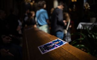 Op een houten kerkbank, waarvoor mensen staan, ligt een folder, met daarop Charlie Kirk, een man in blauw kostuum en zwart haar.