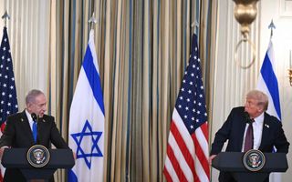 Twee mannen in donkeren pakken staan achter spreekgestoelte met vlaggen in de kleuren blauw, wit en rood op de achtergrond.