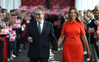 Man in donker pak en vrouw in rode jurk wandelen door een soort erehaag van mensen met Britse vlaggetjes.