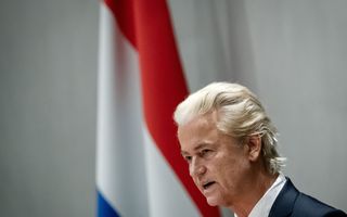 Geert Wilders met geblondeerd haar, rode stropdas en donker pak staat in de Tweede Kamer achter het spreekgestoelte met op de achtergrond de Nederlandse vlag.