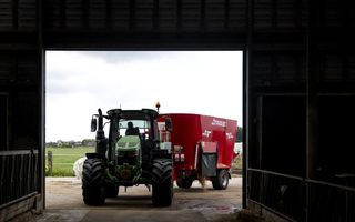 Een groene tractor rijdt met een rode zogeheten voermengwagen een stal binnen. Op de achtergrond zie je weiland.