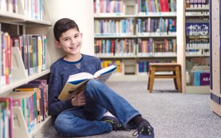Jongen leest zittend op de grond een boek in een bibliotheek.