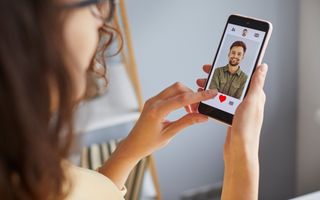 Meisje met bruin haar kijkt op haar telefoon naar een datingapp. Op haar mobieltje is een lachende jongeman zichtbaar.