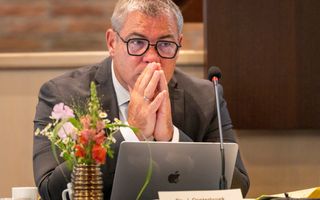 Man met donkere bril achter laptop en achter vergadertafel. Houdt peinzend beide handpalmen tegen elkaar en voor de mond.