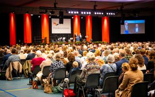 Grote hal vol mensen, zittend op rijen stoelen. Vooraan een podium met twee mannen erop, ook een groot beeldscherm.