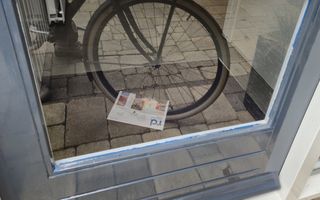Een krant achter een voordeur, op een deurmat. In de spiegeling van het glas van de deur is een fietswiel te zien.