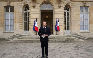 Een man in een donker pak staat achter twee microfoons op een standaard. Achter hem een zandkleurig gebouw, waarvan de deur openstaat. Aan weerszijden van de deur staan een Franse en een Europese vlag.
