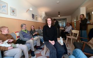Een groep mannen en vrouwen zit of staat in een ruimte met banken en stoelen. In het midden zit een vrouw met krullend haar en zwarte kleding. Op tafel staan koffiemokken.