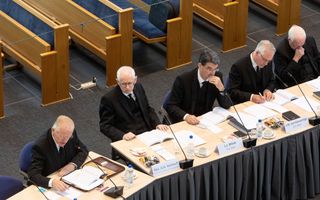 Vijf mannen in zwarte pakken achter vergadertafels.
