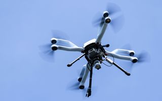 Een witte drone, vier pootjes, vier armen met rotoren en een lichaam met een camera eronder, hangt in een strakblauwe lucht.