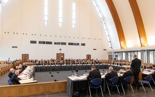 Een modern kerkgebouw met afgevaardigde ambtsdragers in zwarte pakken rond vergadertafels.