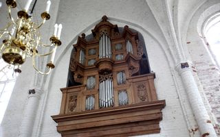 Tegen een wit gepleisterde muur hangt een orgelkas met een eikenhouten kleur. Voor het orgel hangt een grote kroonluchter.