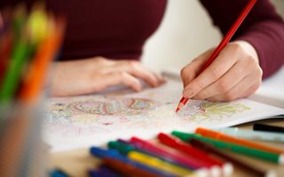 Een opengeslagen kleurboek ligt op tafel. Een hand houdt een rood kleurpotlood vast. Op de voorgrond liggen verschillende kleuren stiften en potloden.