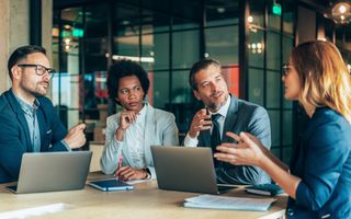 Vier werknemers van een bedrijf, van wie er een donkergetint is, voeren een discussie. Op de tafel twee laptops, agenda’s en documenten.