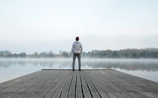 Jonge man staat aan het eind van een aanlegsteiger. Voor hem ligt een meer, met laaghangende mist.