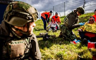 Vrouwelijke militair geknield in het gras bij vrouw met besmeurd gezicht. Op de achtergrond twee mannen bij een man die oogt alsof hij gewond is.